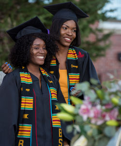 Two BSU graduates in caps, gowns, and sashes, smiling in front of Boyden Hall