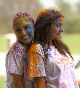 Two students smiling and hugging, their faces and t-shirts splashed with color from the Holi festival