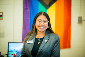 Ellie Cooper standing in front of the pride flag smiling with long straight dark hair wearing silver rimmed eyeglasses and a gray blazer over a black top