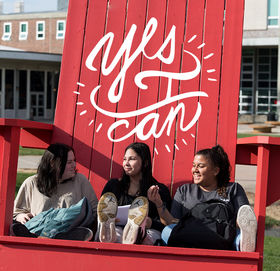 Three students hanging out in the giant red Adirondack chair by Rondileau Student Union. A circle of words above them says, "Yes, I Can."