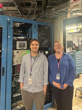 Two people stand in front of a cabinet filled with electronics equipment.