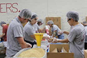 People work together wearing hair nets to pack up meals by tables