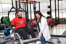 A woman helps show a man how to use a piece of gym equipment