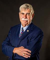 Dr. Michael Vieira smiling with short wavy salt and pepper hair and a gray mustache, wearing a navy blue suit jacket over a light blue button down shirt and a multi color tie