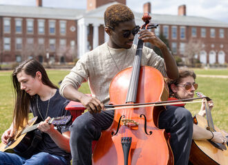 3 male students on Boyden Quad. One sits in a chair playing a cello. The other two sit on the ground playing guitars on either side of the cello player.