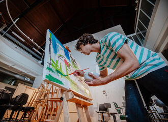 a BSU student stands bending to paint on a large canvas on a wooden easel