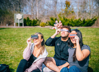 Three students seated on the lawn, watching the solar eclipse through lenses