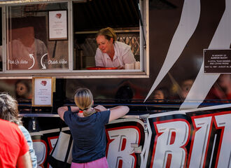 A woman working in the Bear Bites food truck leans out the window smiling while taking an order from a student at the front of the line