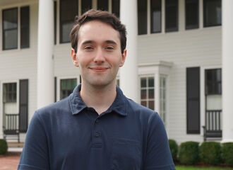 Josh Neff, '27, smiling with short wavy dark brown hair, wearing a navy blue polo shirt and standing in front of the MRISJ building