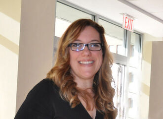 Dr. Melinda Tarsi-Goldfien smiling wearing black rim glasses, long dirty blonde wavy hair and a black v-neck dress