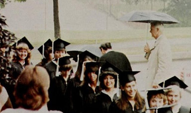 Graduates walk in a line with a man holding an umbrella behind them.