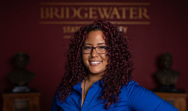 Woman wearing a blue shirt and glasses smiles standing in front of Bridgewater sign