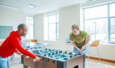 Two men stand next to and play a game of fuseball