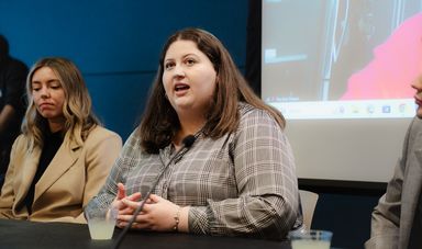 A woman sits at a table with her hands crossed talking to a group of people