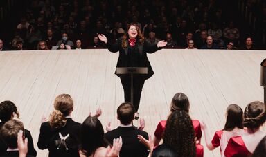 Colleen McDonough opens her arms as she conducts a choir on a stage.