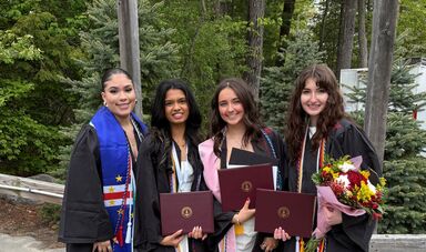 Four female students wearing graduation gowns stand in front of trees.
