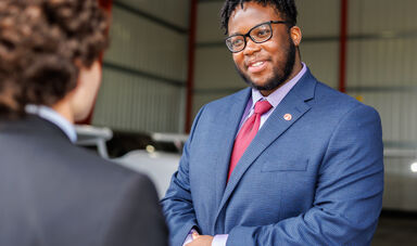 A man wearing glasses smiles as he listens to another man talk