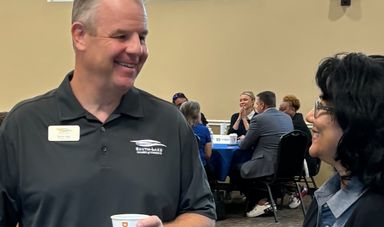 A man stands smiling holding a cup of coffee while talking to a woman who is also miling