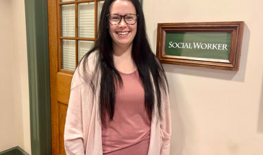 A woman with long brown hair stands smiling next to a social work sign