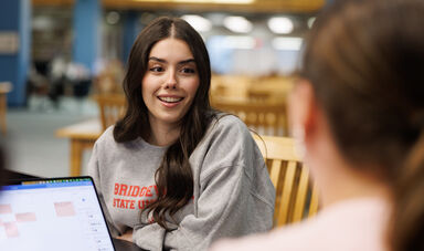 A woman with long brown hair smiles while talking to another woman