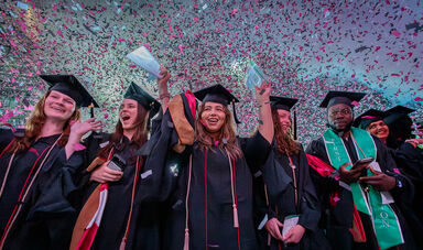 Graduates cheer and raise their hands as confetti rains down.