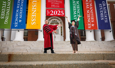 A woman stands on stairs with her hands up in the air, smiling while a man stands next to her also smiling
