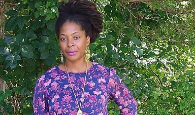 A woman wearing a purple dress stands in front a green tree and poses for camera