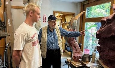 Two men stand next to each other one is pointing to a sculpture while the other looks on