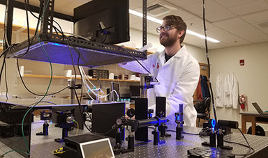 Josh Watts, wearing a white lab coat, looks at a monitor above optical tweezers.