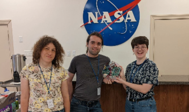 Three people stand holding a circuit board in front of a NASA sign