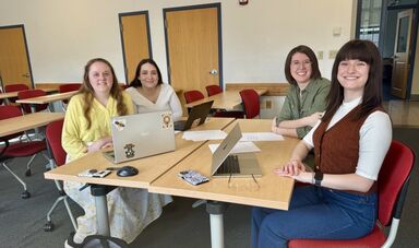 Four people smile while sitting at a table in a classroom.