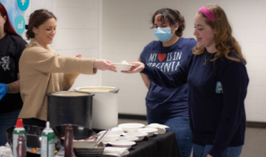 Three people stand around a table and one woman hands a bowl to another