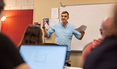 A professor lectures from the front of a classroom as students in the foreground listen.