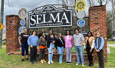 A group of people stand in front of a large sign that reads Selma