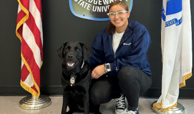 Alysiana Cruz wears a blue jacket and crouches down to pose next to a black K9