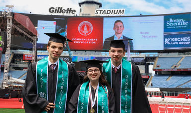 Two men and a woman stand in graduation garb wearing smiles