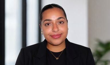 Headshot of Zainab standing in front of windows and a wall.