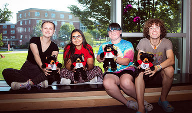 Four students with teddy bears sitting on a window bench in Rondileau Student Union