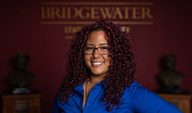 Woman wearing a blue shirt and glasses smiles standing in front of Bridgewater sign 