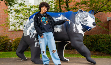 Anaisha stands next to a blue bear statue and points at it smiling 