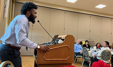 Standing at a podium, Joseph Clark speaks to people seated at tables.