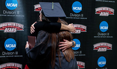 A graduate hugs a coach in front of a BSU athletics backdrop.