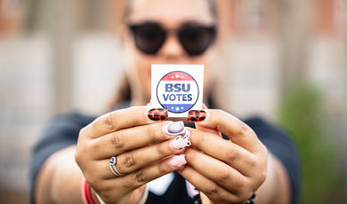 A person is holding a BSU Votes sticker in front of their face with colorful fingernails 