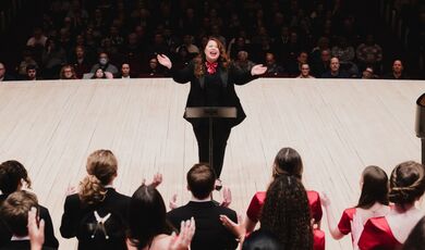 Colleen McDonough opens her arms as she conducts a choir on a stage.