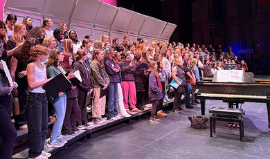 A large group of students sing on a stage next to a piano.