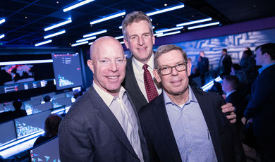 Three men stand together smiling inside of a room with monitors 