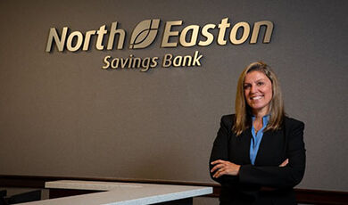 Cate Adams stands in front of a wall with the North Easton Savings Bank logo on it.