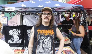 A man with glasses wearing a hat stands in front of a tent selling tshirts 