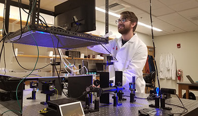 Josh Watts, wearing a white lab coat, looks at a monitor above optical tweezers.