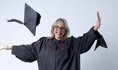 Lesly Freed, wearing her black graduation gown, tosses her cap in the air in front of a white background.